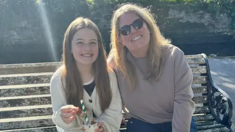 Sam Marlow Rubie is sitting on a wooden bench with long brown hair and a cream cardigan. She is holding a drink with a green and white straw and is sitting beside her mum Sam smiling. Sam is wearing a light purple jumper and has long blonde hair and sunglasses.