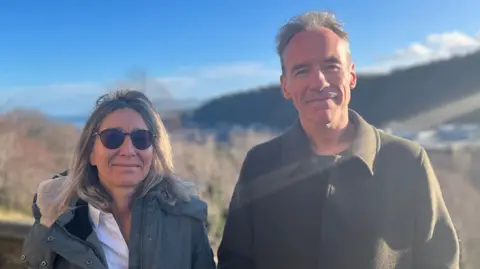 Prof McStay and Prof Bakir are standing outside and both looking at the camera  with hillsides behind them.  Prof Bakir has shoulder-length hair and sunglasses. She is wearing a grey winter coat. Prof McStay has short grey hair and is wearing a green wool coat.