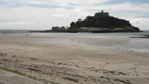 BBC A view of St Michael's Mount from Marazion. The beach is sandy and the tide is out. The island has a castle situated at the top.