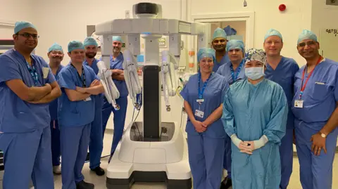 UHNM A group of people dressed in blue hospital scrubs, standing around a white piece of robotic equipment. They are standing in a light-coloured room, which appears to be a hospital setting.