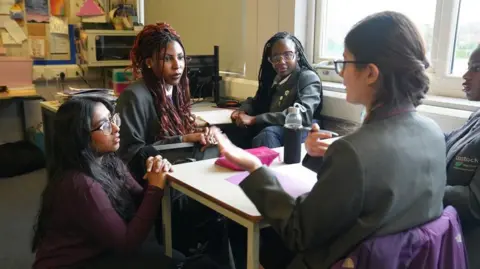 Ann Gannon / BBC A teacher, with long back hair and glasses kneels down beside a school desk talking to some students . The students are wearing black blazers and white shirts.