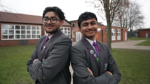 Ann Gannon / BBC Abdullah and Bakhtiar are standing back to back with their arms folded smiling at the camera. They are wearing their gray school blazers, purple ties and white shirts.